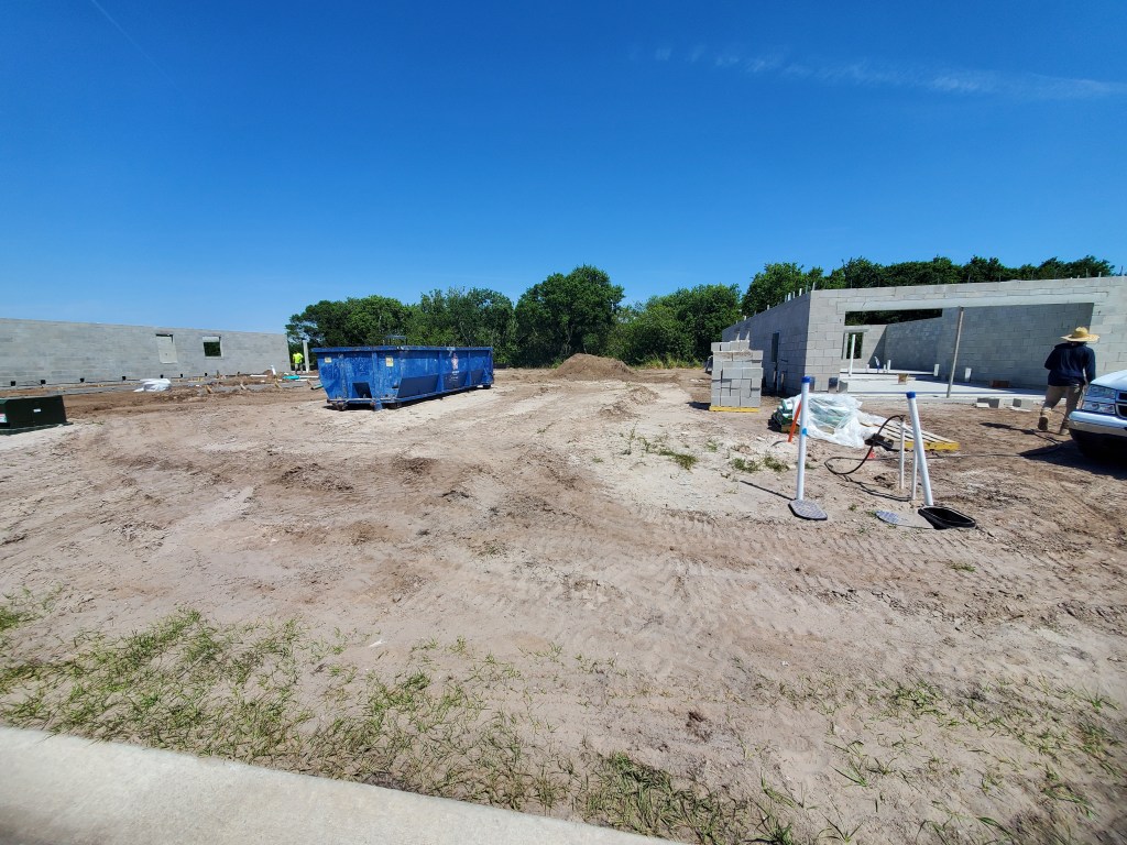 Construction site with partially built concrete walls, a blue dumpster, scattered materials, and dirt ground under a clear blue sky. Trees and greenery are visible in the background—a scene often visited by Rich Noto Realtor for project insights.